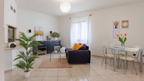 Un salon moderne comprenant un canapé, une table à manger et des chaises, un coin cuisine avec des placards et une fenêtre, ainsi qu’une plante en pot.