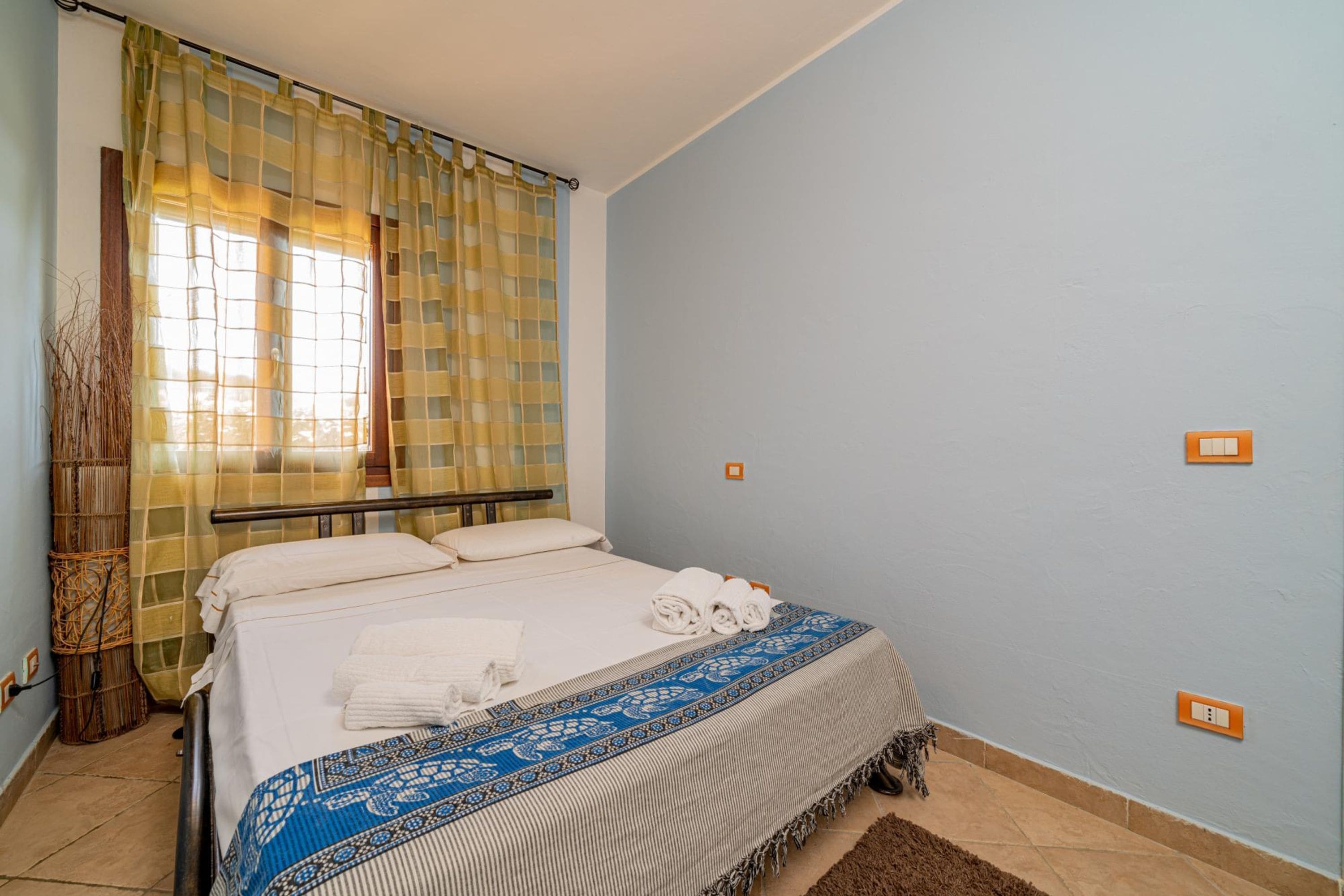 Een net opgemaakt bed met een blauw gebedrukte sprei, een raam met geel geruit gordijn en een stopcontact aan de rechterkant.
