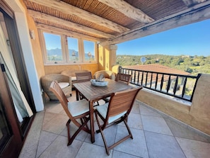 Un balcón con mesa y sillas de madera, vista al campo y piso de baldosas.