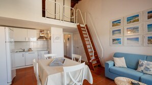 A kitchen with white cabinets, a dining table with chairs, a blue sofa, a wicker side table, and a wooden staircase. - Casa Petralana by Wonderful Italy (Olbia)