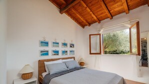 A bedroom with a wooden ceiling, a bed with grey bedding, a nightstand with a lamp, a window with a view of trees, and several framed pictures on the wall. - Casa Petralana by Wonderful Italy (Olbia)