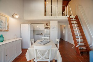 Una cocina con armarios blancos, una mesa de comedor con sillas, un refrigerador y una escalera que sube a un nivel superior.
