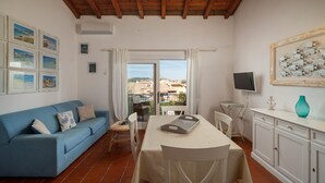 A living room with a blue sofa, a dining table with chairs, a TV, and a view of buildings through a window. - Casa Petralana by Wonderful Italy (Olbia)