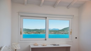 A bedroom with a bed, a nightstand, a desk, and a window offering a view of a coastal landscape. - Casa Azzurra by Wonderful Italy (Olbia)