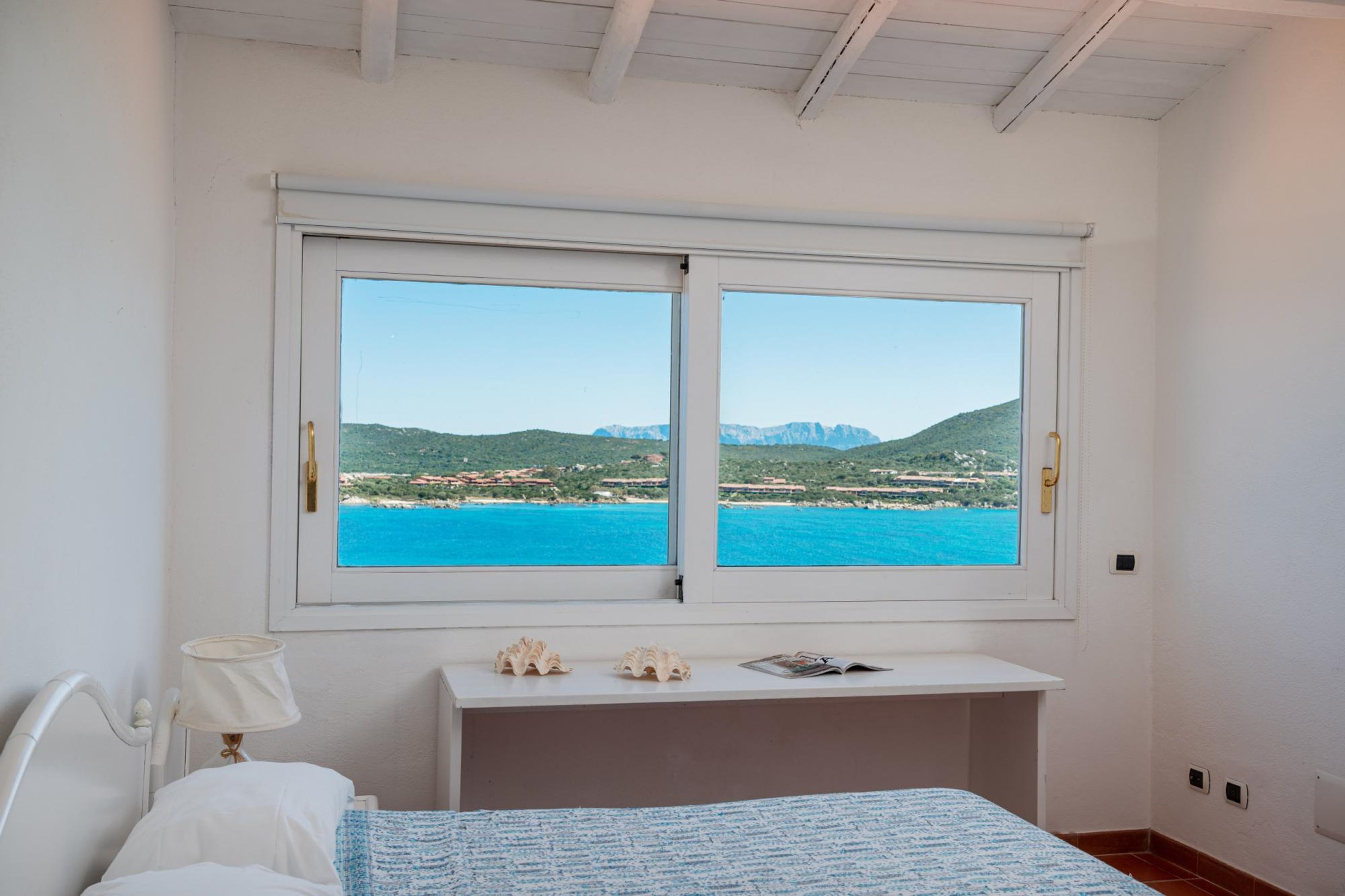 A bedroom with a bed, a nightstand, a lamp, a desk with decorative items, and a window with a view of a coastal landscape.
