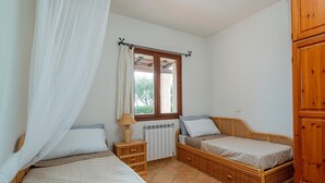 A bedroom with two single beds, a wooden wardrobe, a bedside table with a lamp, and a window with a curtain. - Casa Helen by Wonderful Italy (Olbia)