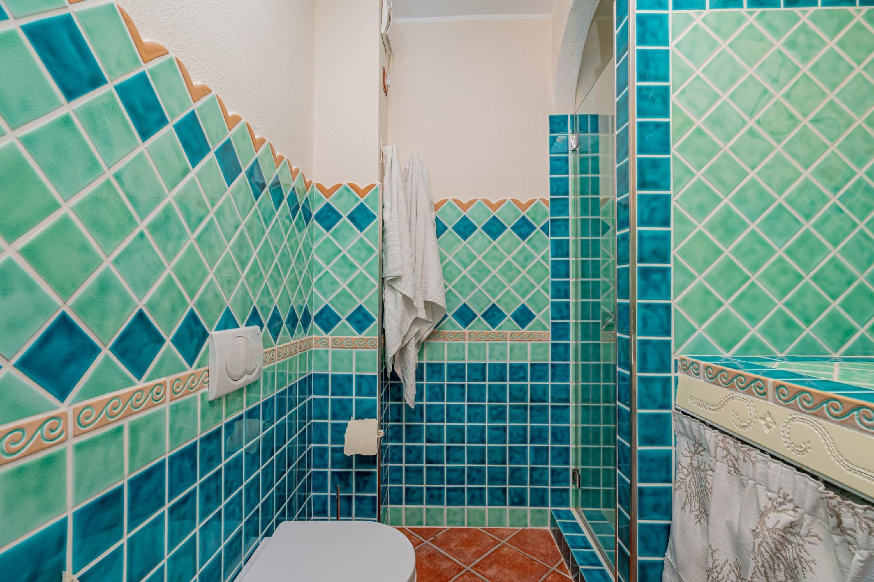 Un baño con mosaicos azules y verdes, un inodoro blanco y una toalla colgada en la pared.
