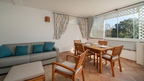 A room with a grey sofa, teal cushions, wooden dining table with chairs, and a window with a lattice design. - Sweet Mavy Incantevole Dimora by Wonderful Italy (Olbia)
