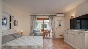 A bedroom with a bed, a TV, a dining area with chairs and a table, and a balcony with curtains. - Sweet Mavy Incantevole Dimora by Wonderful Italy (Olbia)