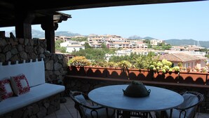 A terrace with a stone wall, a white cushioned bench, a round table with chairs, and a view of a hillside town.