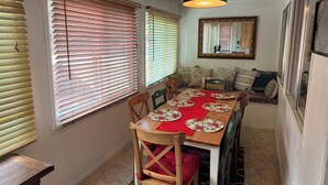 A dining area with a wooden table set for four, flanked by two windows with blinds, a ceiling fan, and a mirror on the wall.
