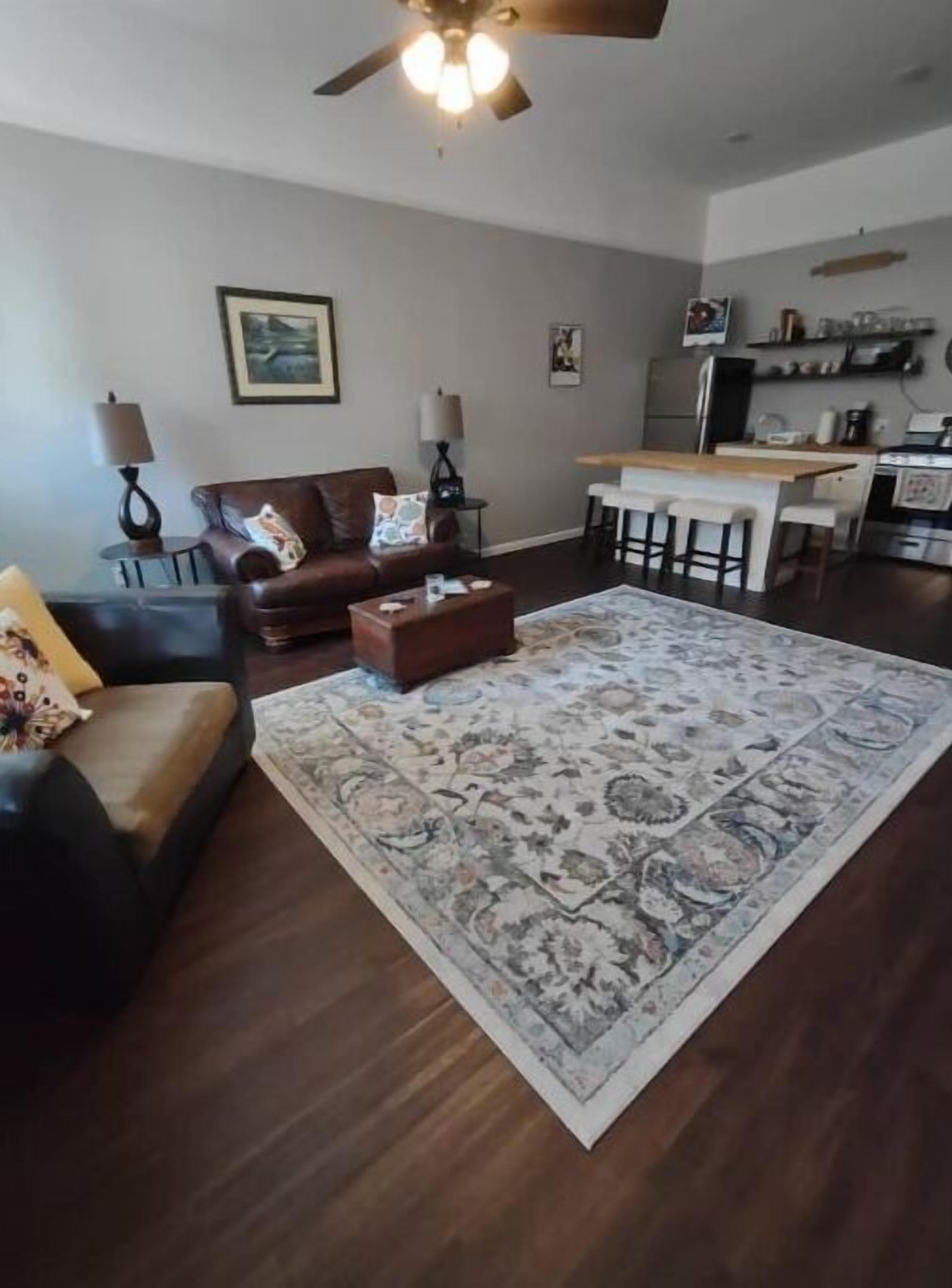 Ein Wohnzimmer mit dunklem Holzboden, einem braunen Ledersofa, einem Couchtisch und einem gemusterten Teppich. Es gibt einen Deckenventilator, einen Essbereich mit einer Bar und Küchengeräte im Hintergrund.