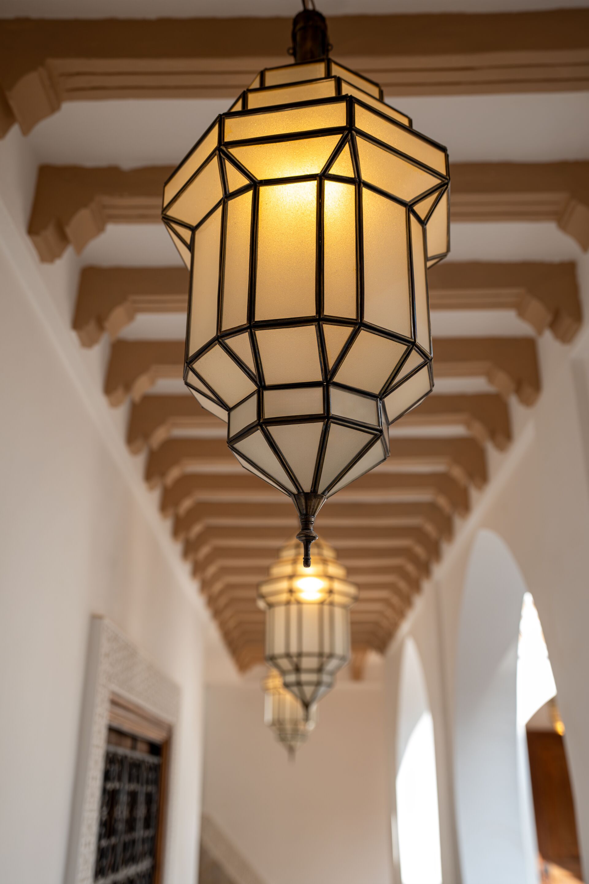 A geometric pendant light fixture in a hallway with white walls and wooden beams.