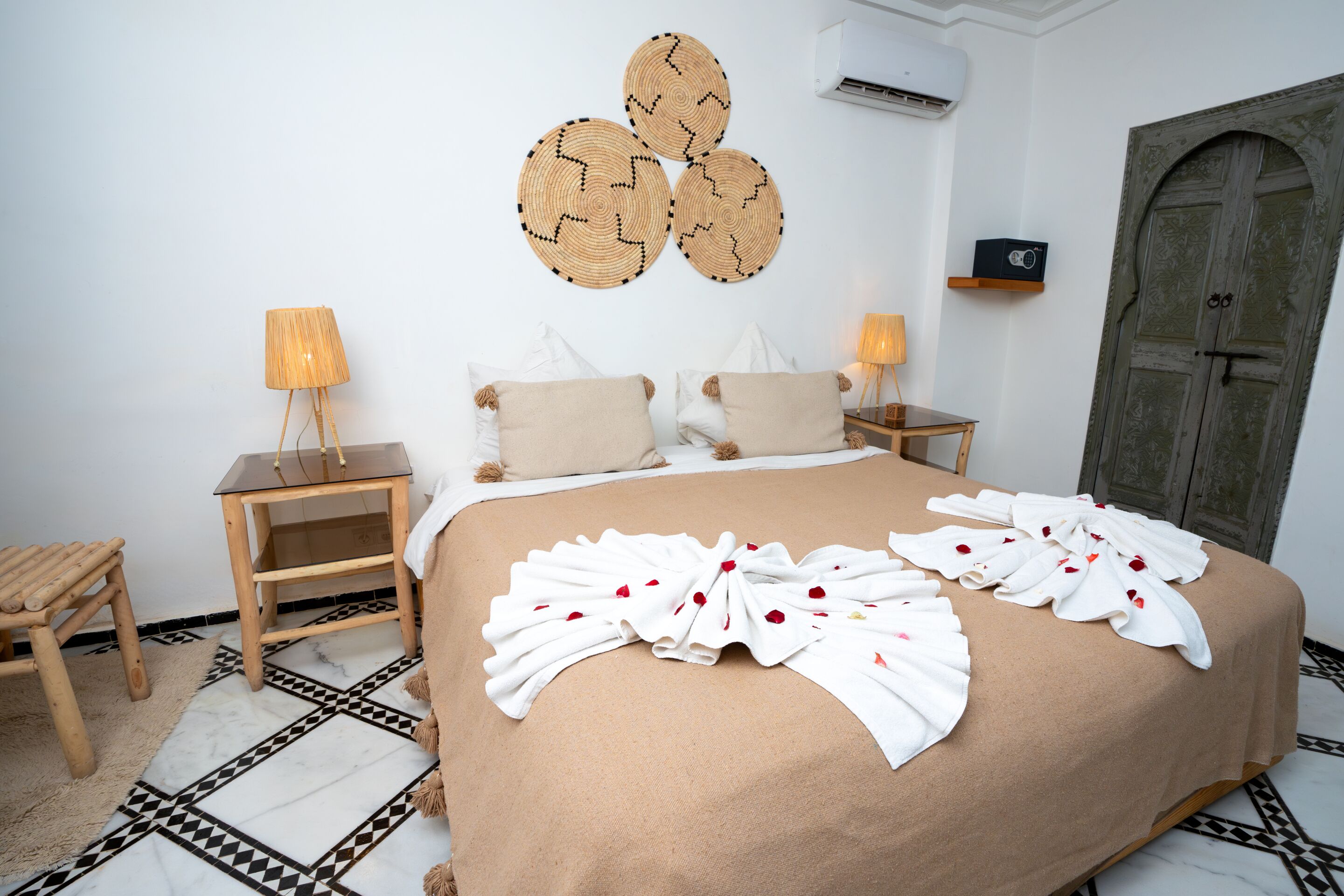 A neatly made bed with a brown bedspread and white towels arranged in a fan shape, flanked by two bedside tables with lamps, a wooden stool, and a wall-mounted air conditioner.