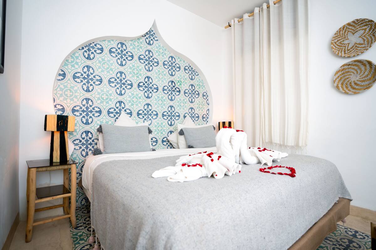 A bedroom with a patterned headboard, a bedside table with a lamp, and a bed with a grey blanket and red accents.