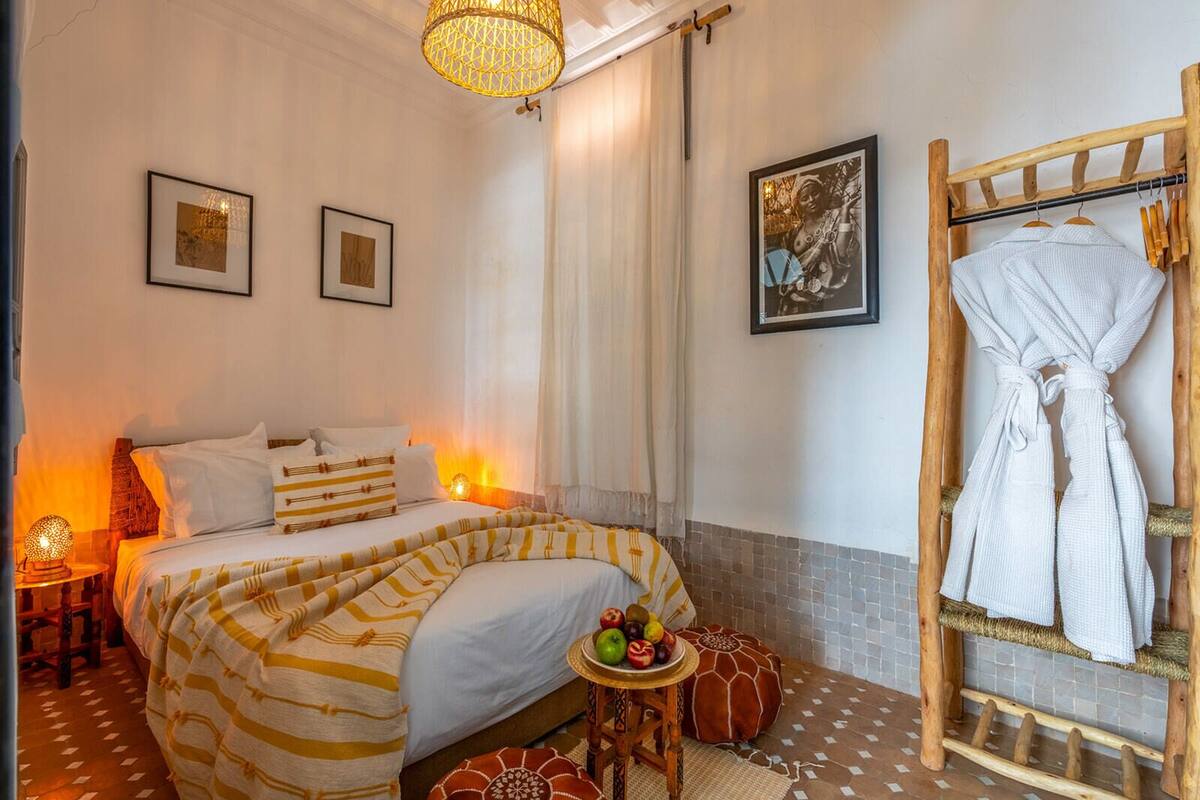 A hotel room with a bed, bedside lamps, a wooden rack with robes, a small table with fruit, and a wall-mounted picture.
