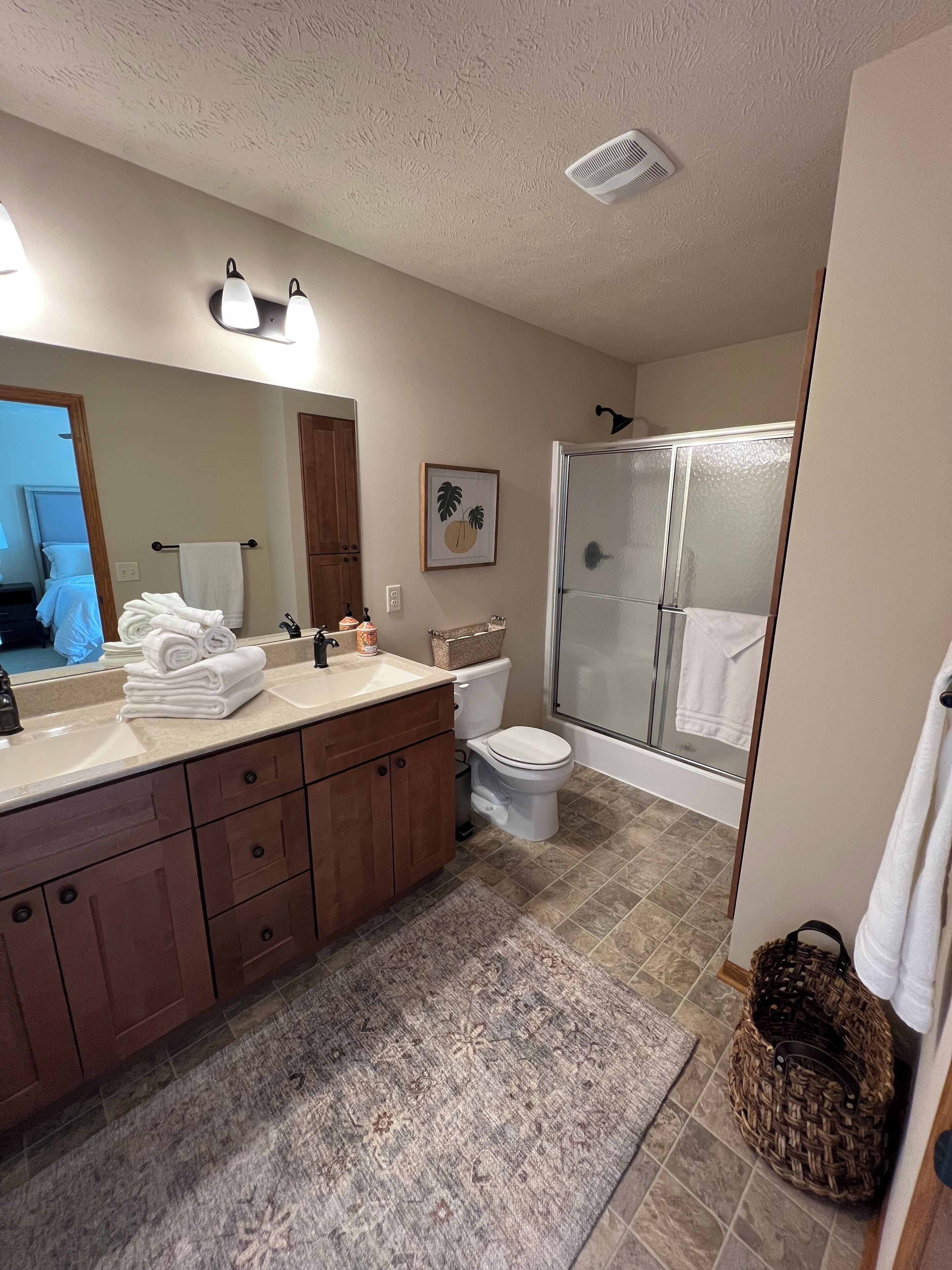 Een badkamer met een grote spiegel, een wastafel met handdoekhouder, een toilet, een douche met glazen deur en een vloerkleed.