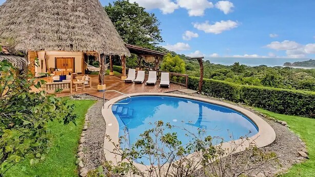 Une maison à toit de chaume avec une piscine et des chaises longues.