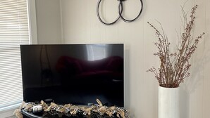 Ein Wohnzimmer mit einem Flachbildfernseher auf einem schwarzen Standfuß, einer hohen Vase mit Zweigen und einer kreisförmigen Wanddekoration.