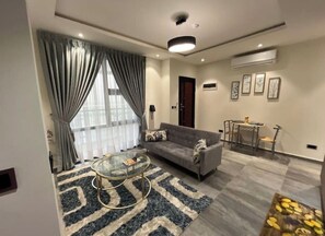 A modern living room with a sofa, a glass coffee table, and a dining area with a small table and chairs. - AP1 Park Lux Apartments  (Accra)
