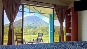 A room with a bed, two chairs, and a table. It features a large window with curtains, offering a view of a mountain. - Villa Luna del Arenal (La Fortuna)