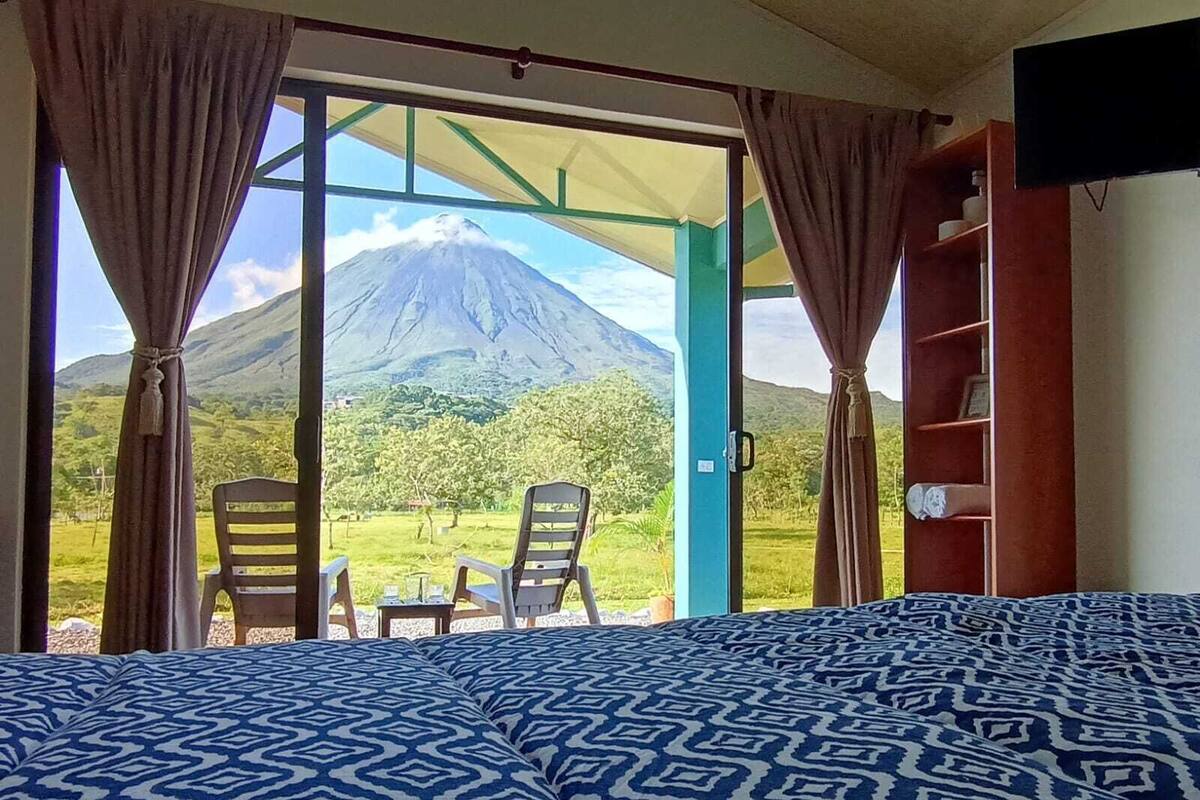 A room with a bed, two chairs, and a table. It features a large window with curtains, offering a view of a mountain.