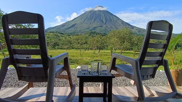 Two rocking chairs facing a mountain with a view of a grassy landscape and a table with glasses and a vase. - Villa Luna del Arenal (La Fortuna)