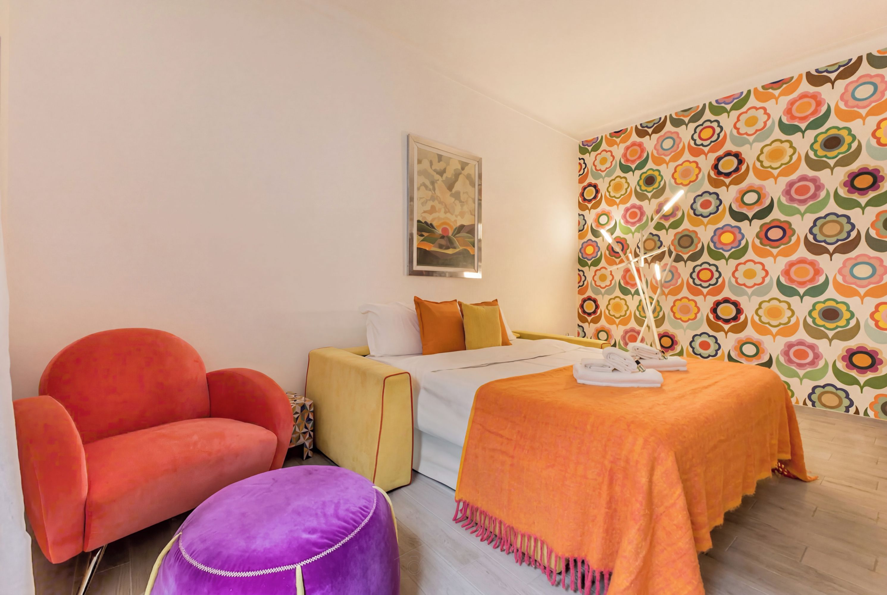 A hotel room with a bed, a chair, a small table, and a wall with a colorful floral pattern.