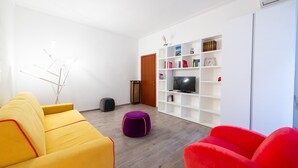 A modern living room with a yellow sofa, a red armchair, and a purple ottoman. There is a white shelving unit with books and a television. - Casa Riboty - One Bedroom Apartment, Sleeps 2 (Rome)