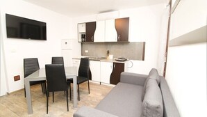 Une cuisine moderne avec des meubles blancs, un four encastré et une machine à café. On y trouve également un coin repas avec un canapé gris et deux chaises noires.