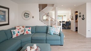 Una sala de estar con un sofá seccional color aguamarina, un espejo redondo y almohadones con motivos náuticos. Una escalera conduce a un nivel superior.