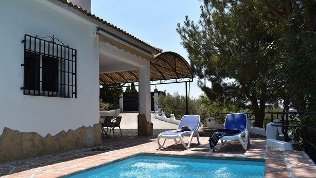 A swimming pool with two lounge chairs, a covered patio, and a stone wall.