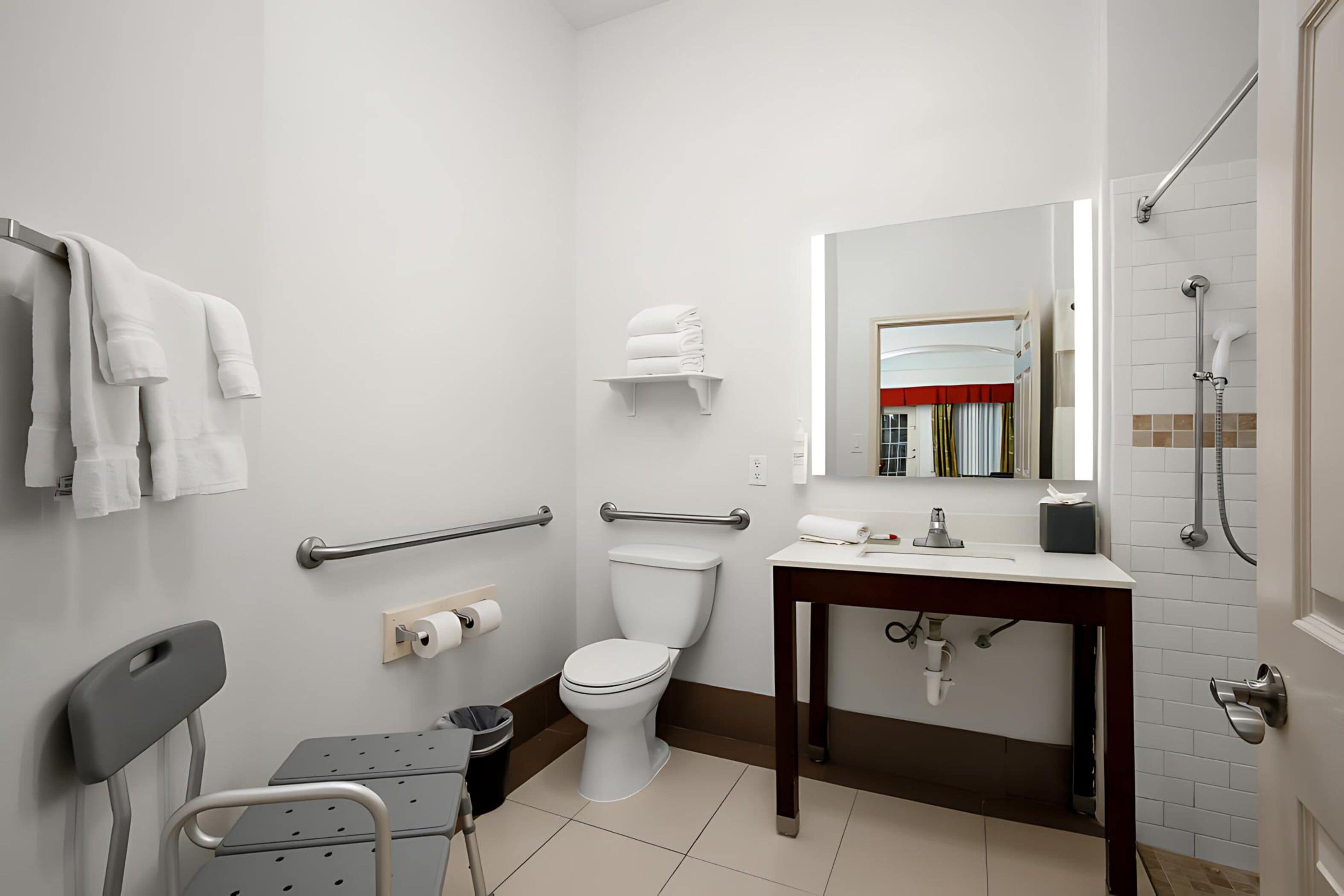 Un baño moderno con inodoro, lavabo y ducha. Incluye una silla, barras de apoyo y un espejo.