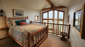 A bedroom with a large bed, wooden headboard, a painting on the wall, and a view through a window.