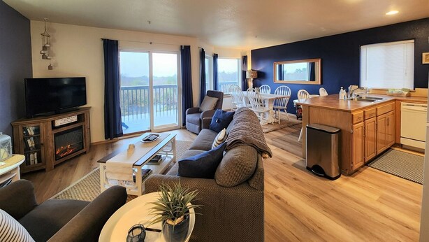 A spacious living area with a sofa, TV, and fireplace, a dining area with chairs, and a kitchen with a sink and cabinets. - Windsong # 6 Beautiful condo with partial ocean view (Ocean Shores)