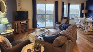 A living room with a television, sofa, armchair, dining table, and a balcony with curtains. - Windsong # 6 Beautiful condo with partial ocean view (Ocean Shores)