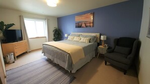 A bedroom with a bed, a chair, a nightstand, a television on a dresser, and a window with curtains. - Windsong # 6 Beautiful condo with partial ocean view (Ocean Shores)