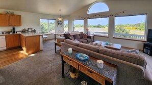 A spacious living area with a sofa, a dining table, and a kitchen area with wooden cabinets. Large windows provide views of the outdoors.