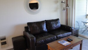 A living room with a black leather sofa, a wooden coffee table, a wall-mounted TV, and a sliding glass door leading to an outdoor area. - Nautilus # 202 - Ocean Front Condos (Ocean Shores)