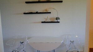A room with a white table and two clear plastic chairs. There are three black shelves on the wall, each holding different items. - Nautilus # 202 - Ocean Front Condos (Ocean Shores)