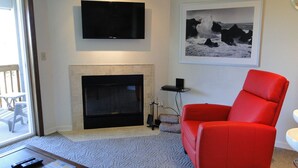 A living room with a fireplace, a red armchair, a flat-screen TV, and a framed photograph on the wall. - Nautilus # 202 - Ocean Front Condos (Ocean Shores)