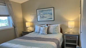 Une chambre à coucher bien rangée, avec un grand lit, deux tables de chevet avec des lampes, un tableau encadré au mur et une fenêtre avec des rideaux.