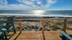 Une terrasse en bois avec des chaises, donnant sur une plage où l’on peut admirer les vagues et un ciel dégagé.