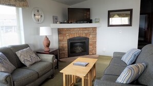 A living room with two grey sofas, a wooden coffee table, a fireplace, a clock, and a television mounted above the fireplace.
