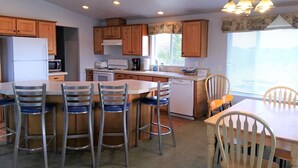A compact kitchen with wooden cabinets, a refrigerator, microwave, stove, and a sink. There are four bar stools and a dining table with chairs.
