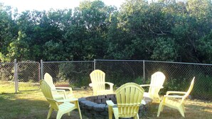 Un grupo de sillas Adirondack amarillas dispuestas alrededor de un brasero de piedra en un patio césped, con una valla de malla y árboles al fondo.