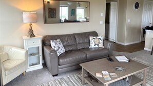 A living room with a brown leather sofa, a white armchair, a coffee table with books and remote controls, a striped lamp, and a mirror with a 'BEACH HOUSE' sign above it. - Bristol Court C- Beautiful 3 Bedroom (Ocean Shores)
