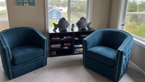 A room with two blue armchairs, a bookshelf, decorative fish sculptures, and a window with a view of a house and greenery. - Bristol Court C- Beautiful 3 Bedroom (Ocean Shores)