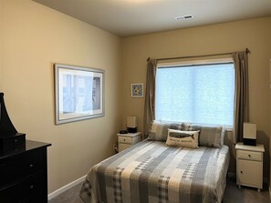 A bedroom with a bed, a nightstand, a dresser, a window with curtains, and a framed picture on the wall.