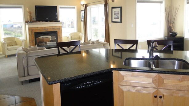 A modern kitchen with a central island, built-in dishwasher, and a dining area with a fireplace. - Bristol Court D- Beautiful 3 Bedroom (Ocean Shores)
